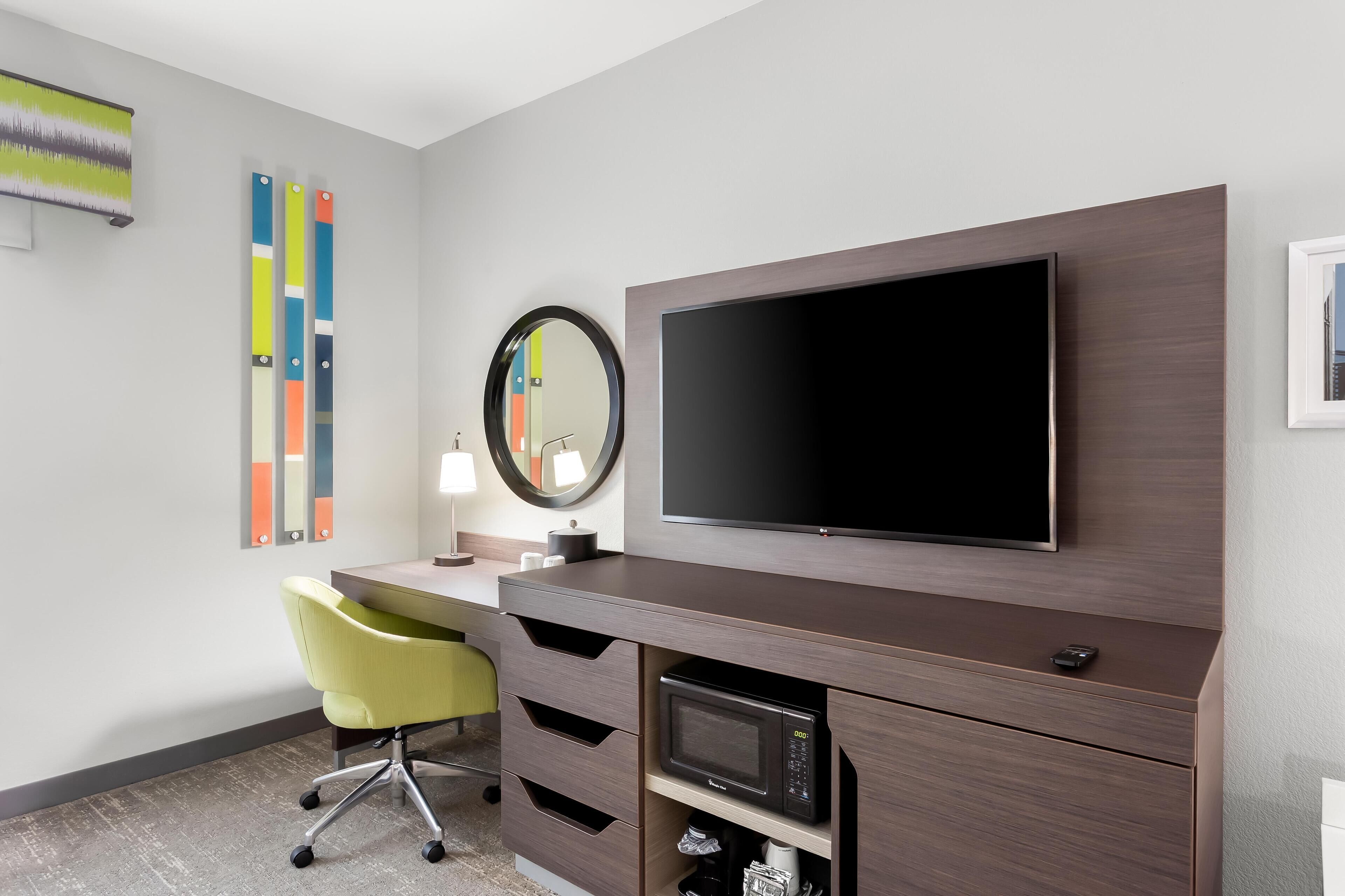 A modern hotel room with a flat-screen TV mounted on the wall, a desk with a chair, a microwave, and a coffee maker.