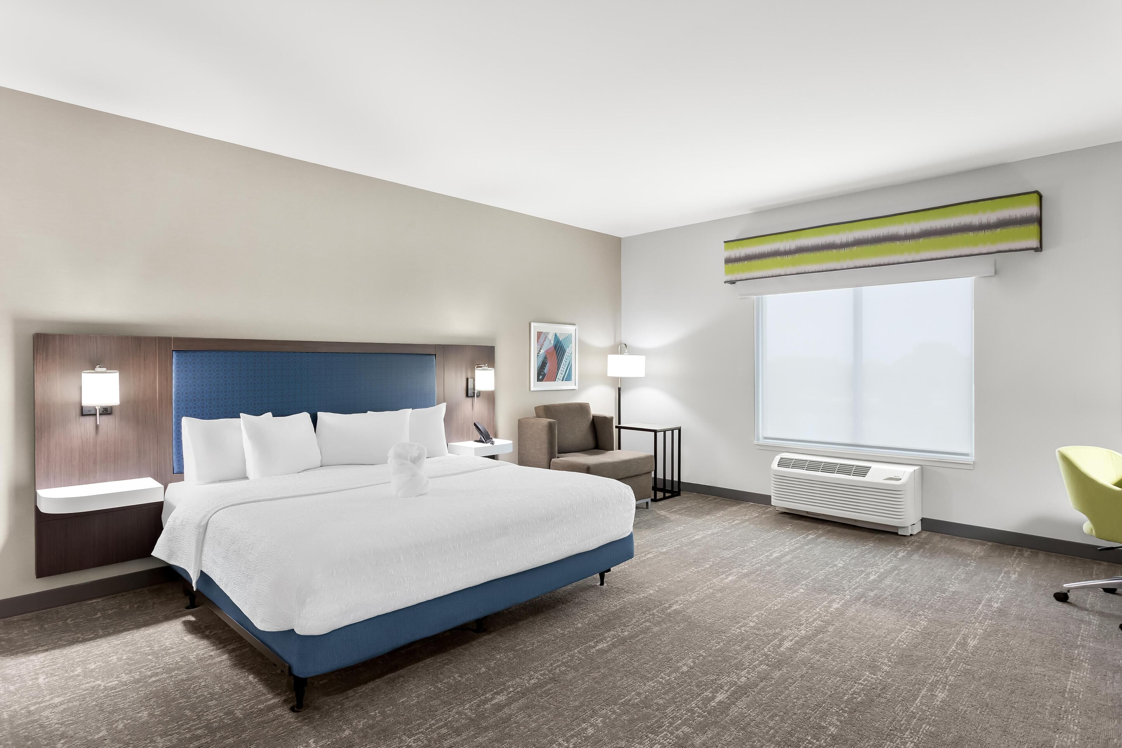 Una habitación de hotel moderna con una cama grande, mesitas de noche, un escritorio, una silla, un ventanal y un aire acondicionado instalado en la pared.
