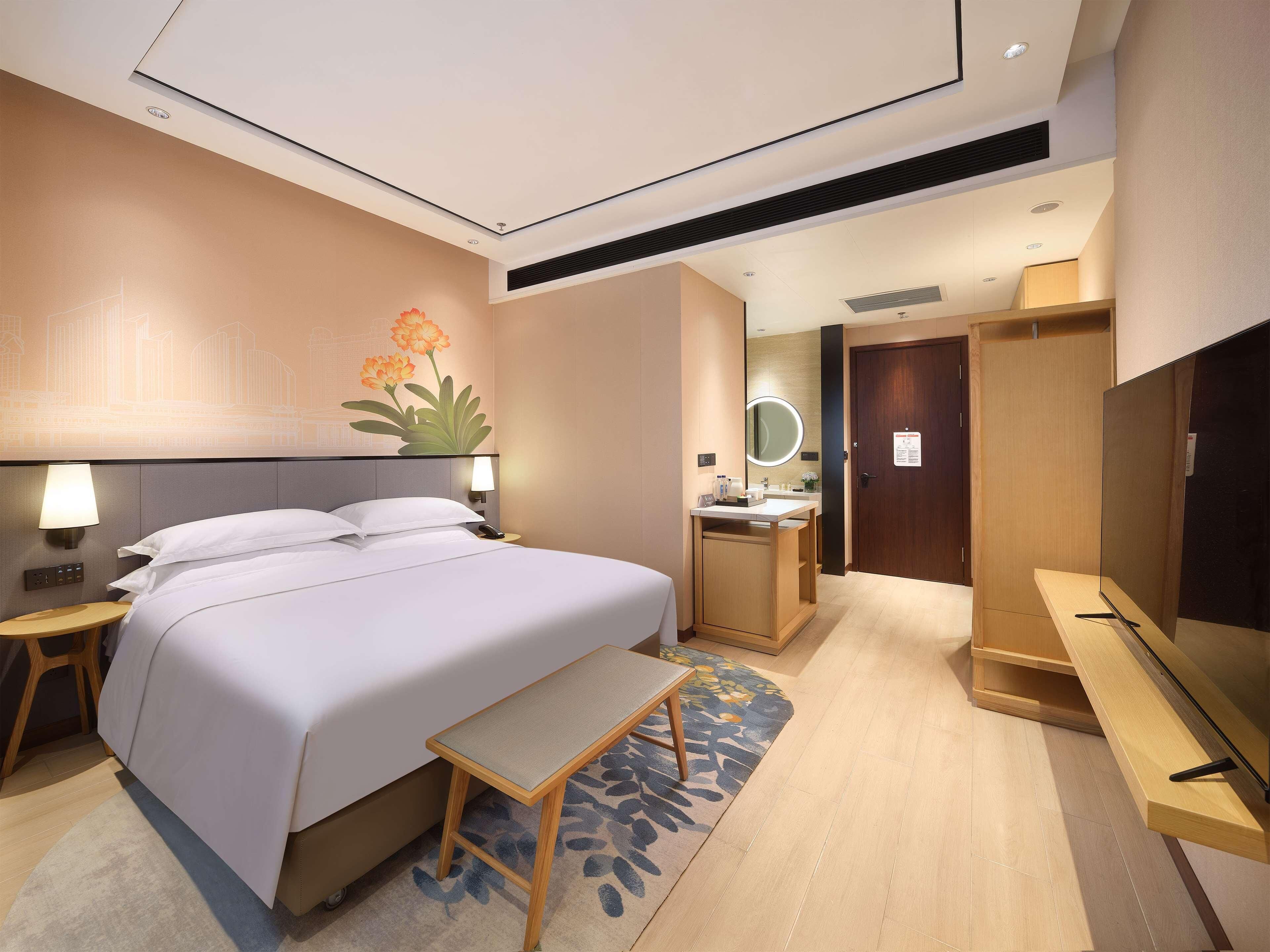 A hotel room with a bed, bedside table, lamp, mirror, vanity, and a TV.