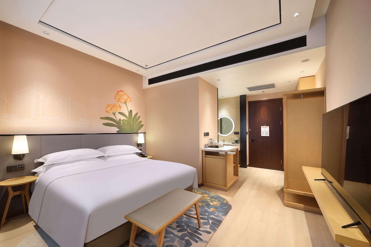 A hotel room with a bed, bedside table, lamp, mirror, vanity, and a TV.