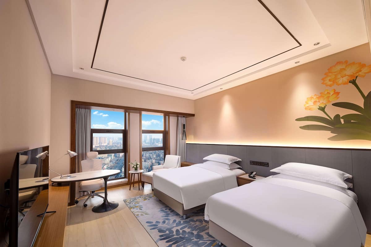 A hotel room with two beds, a desk, a chair, and a view of the city.
