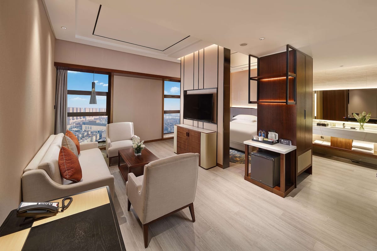 A modern hotel room with a sofa, armchair, coffee table, TV, and a small kitchenette.