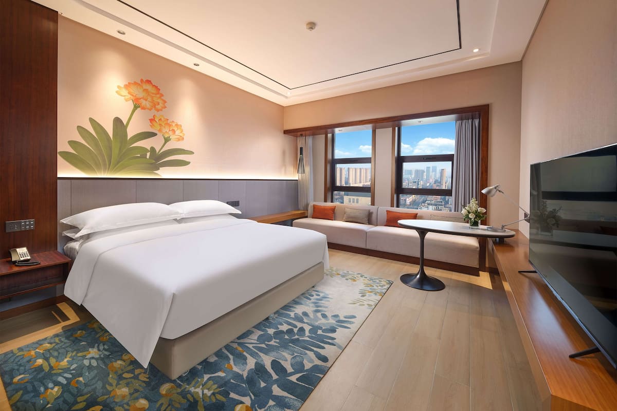 A modern hotel room with a large bed, a sofa, a small table, a TV, and a city view through large windows.