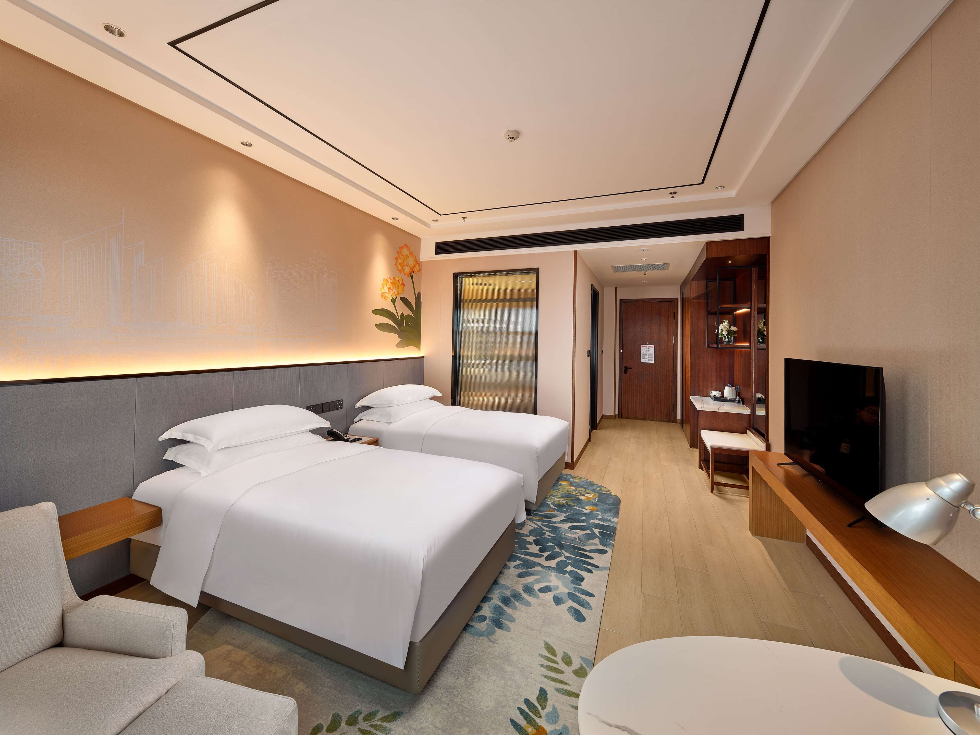 A modern hotel room with two beds, a desk, a TV, and a seating area.