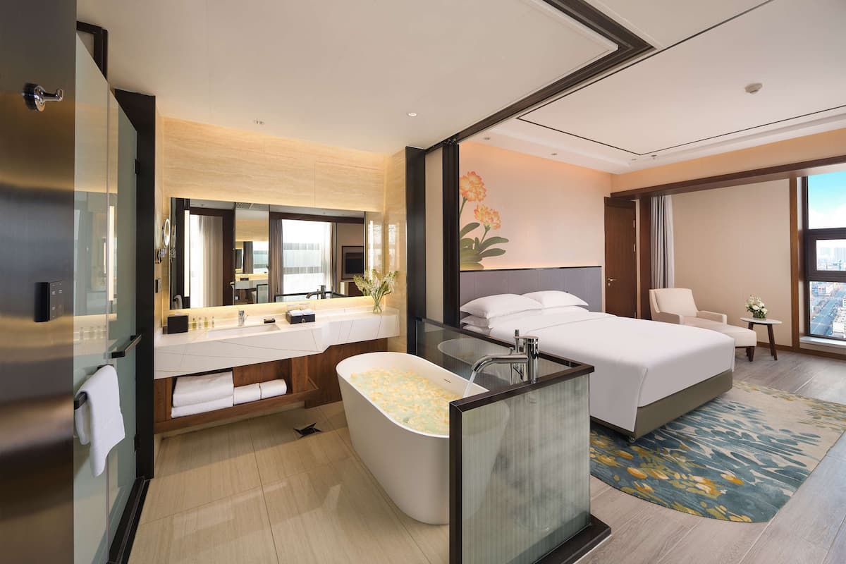 A modern hotel room with a large bed, a separate bathroom with a bathtub, a vanity with a mirror, and a large window offering a city view.