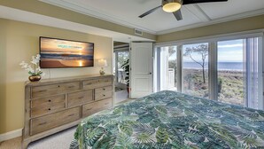 Une chambre avec un lit, un ventilateur de plafond, un téléviseur fixé au mur, une commode et une vue sur l’extérieur grâce à de grandes fenêtres.