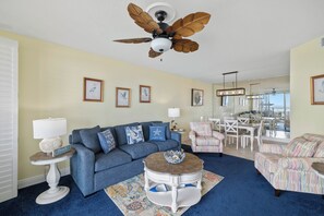 A living room with a blue sofa, a round coffee table, a ceiling fan, and framed pictures on the wall.