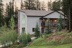 A modern two-story house with a stone facade, a balcony, and a view of a forested hillside.