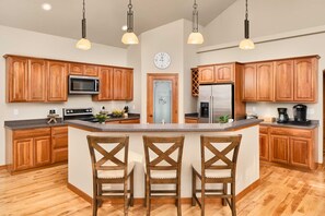 A kitchen with wooden cabinets, a central island with chairs, a microwave, a refrigerator, and pendant lighting.