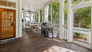 Eine überdachte Terrasse mit Grill, Gartenmöbeln und großen Fenstern.