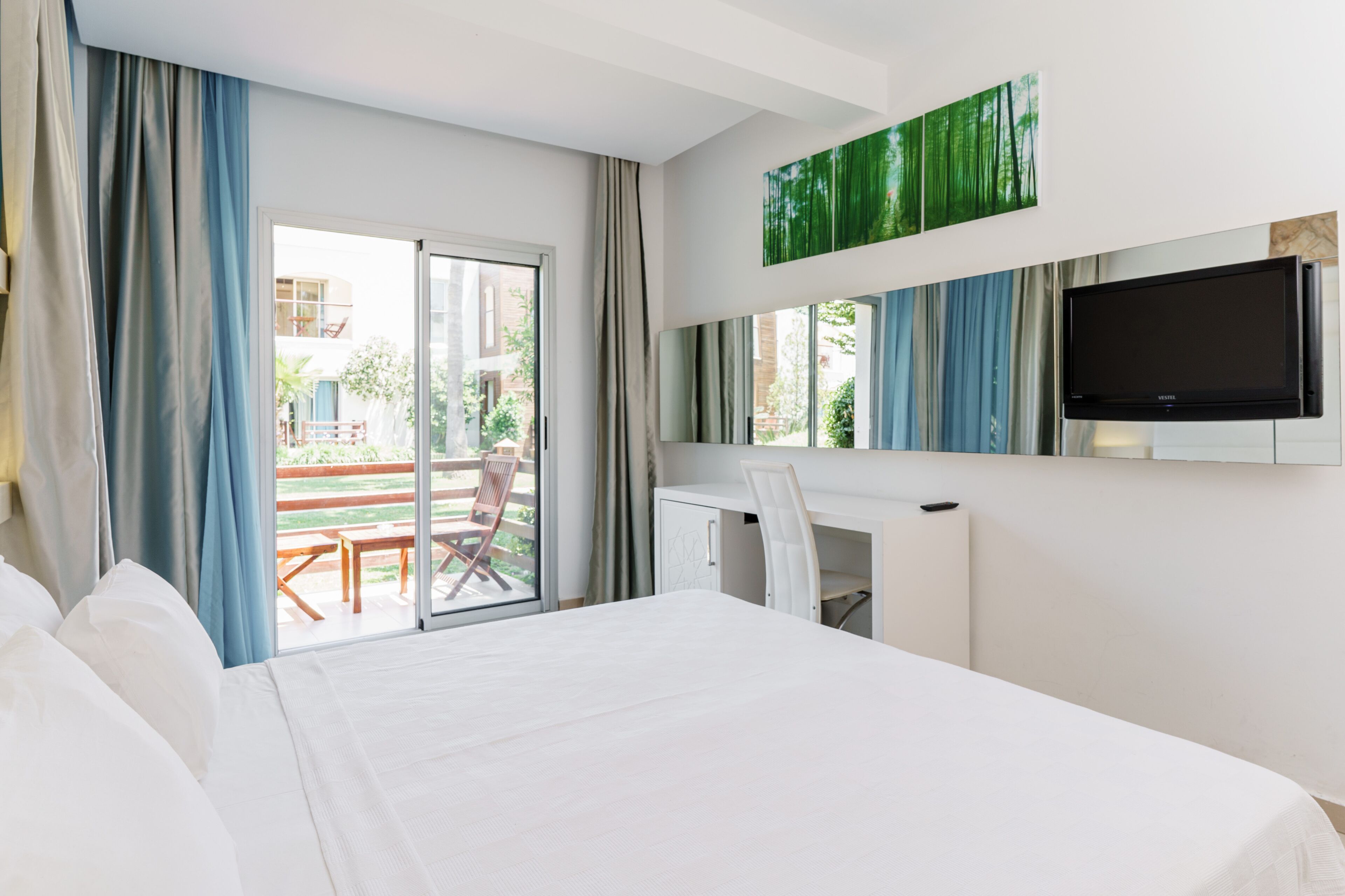 A hotel room with a bed, a desk, a chair, a TV, and a balcony with a view of the outside.