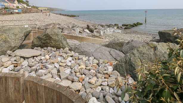 Een rotsachtig strand met een houten looppad naar de zee, huizen op de achtergrond en een heldere lucht.