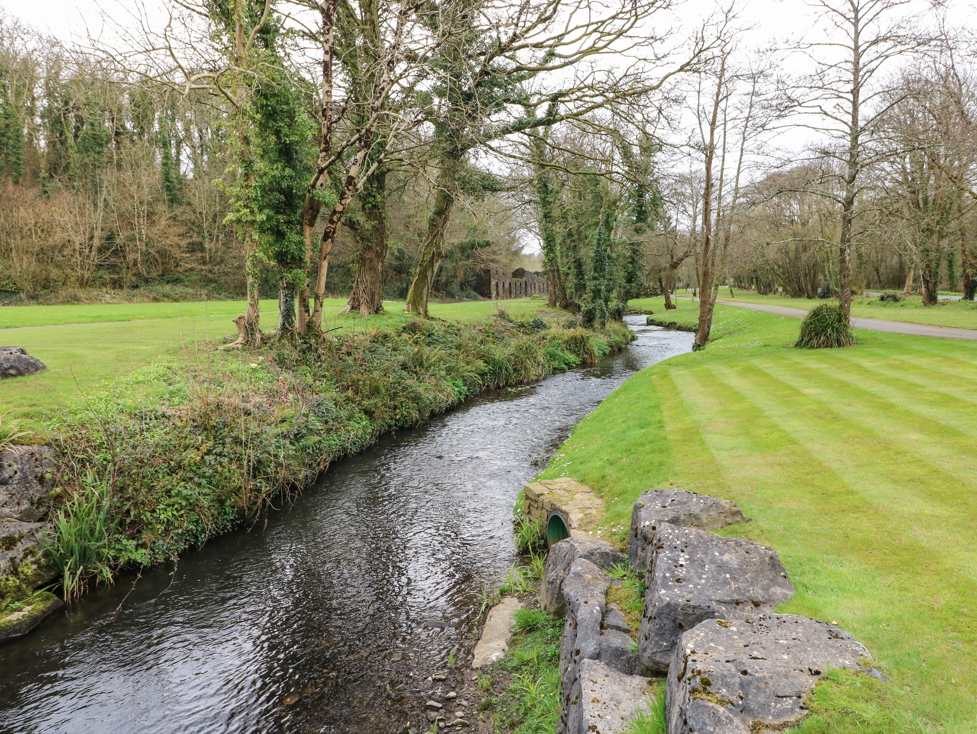 A serene riverside pathway with a narrow stream, surrounded by lush greenery and trees.