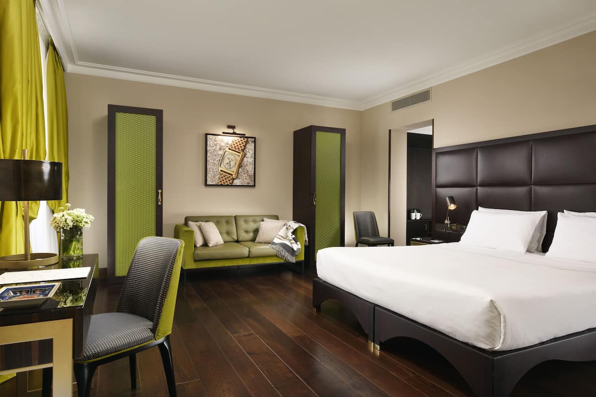 A hotel room with a large bed, a green sofa, a desk with a chair, and a painting on the wall.