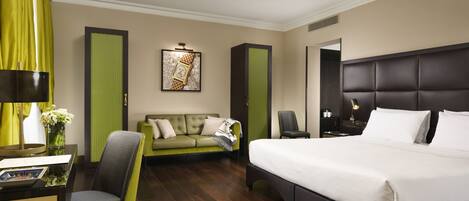 A hotel room with a large bed, a green sofa, a desk with a chair, and a painting on the wall.