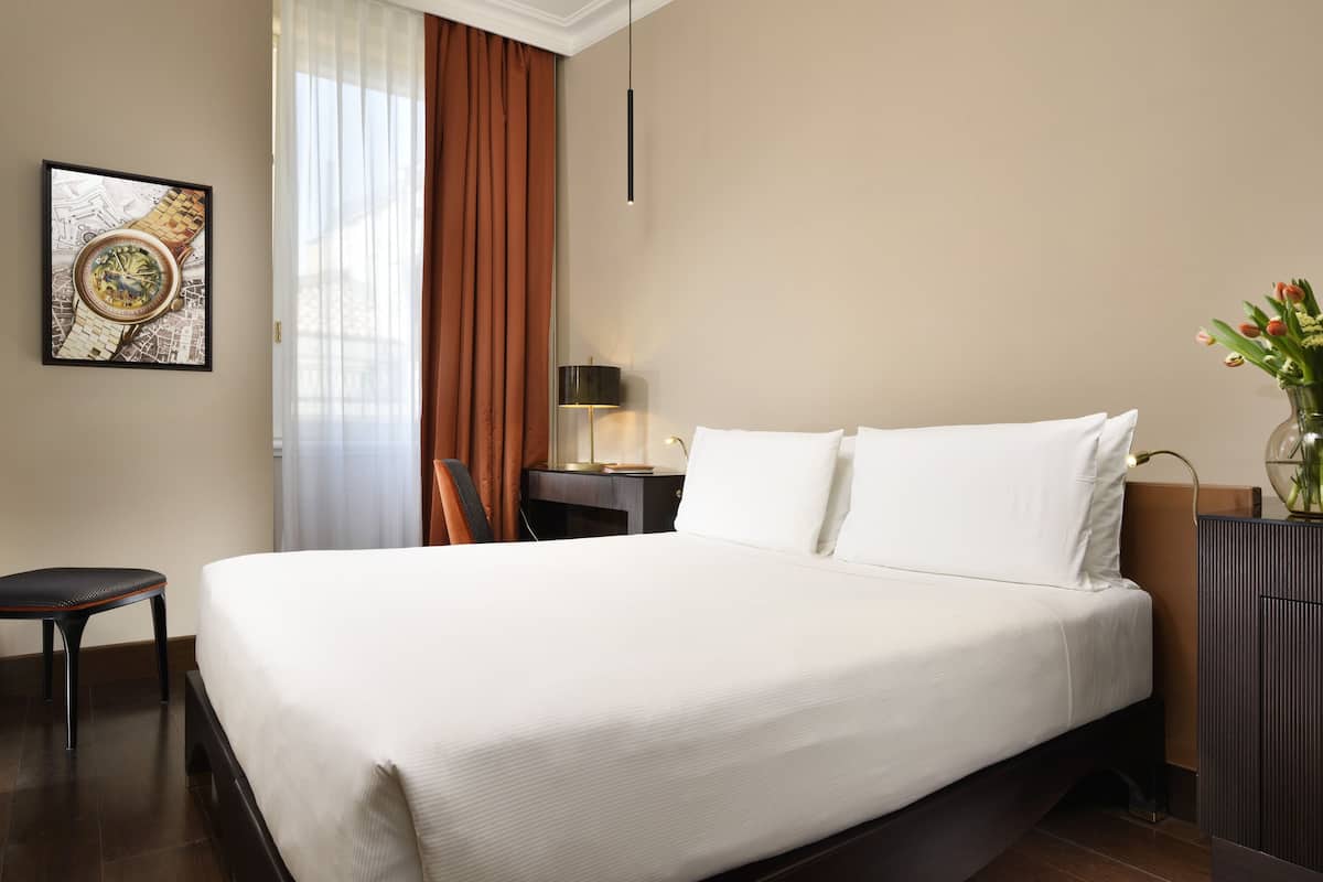 A hotel room with a large bed, a desk with a lamp, a chair, a framed picture on the wall, and a vase with flowers on a cabinet.