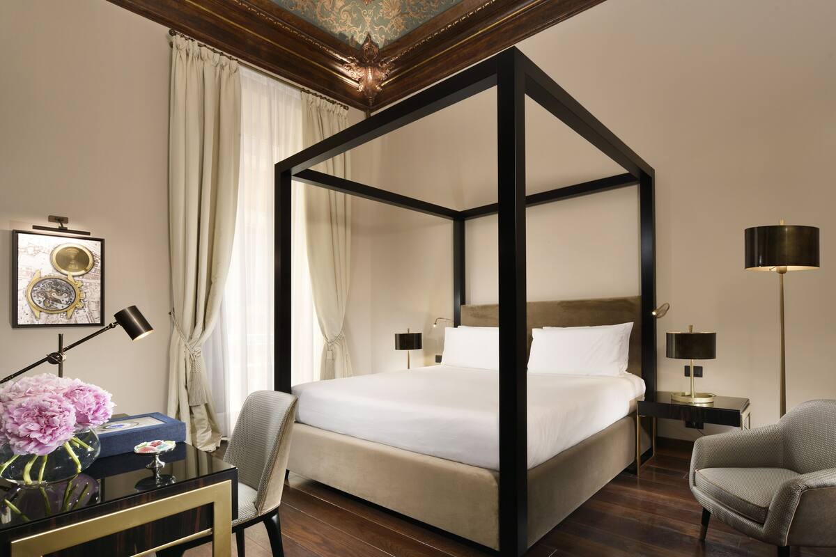 A four-poster bed with a wooden frame, a bedside table with a lamp, a chair, and a desk with a lamp.