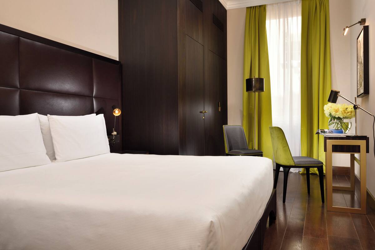 A hotel room with a large bed, a desk with a chair, a lamp, and a vase with flowers.