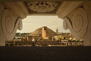 A view through a stone archway with a balustrade, overlooking a historic building with a dome and a tower.
