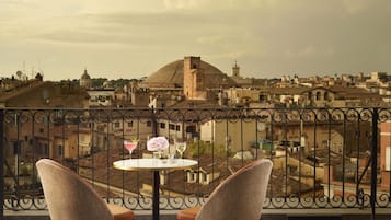 Un balcón con dos sillas de mimbre y una mesita redonda, con vistas a un paisaje urbano histórico con edificios de cúpulas y una torre.