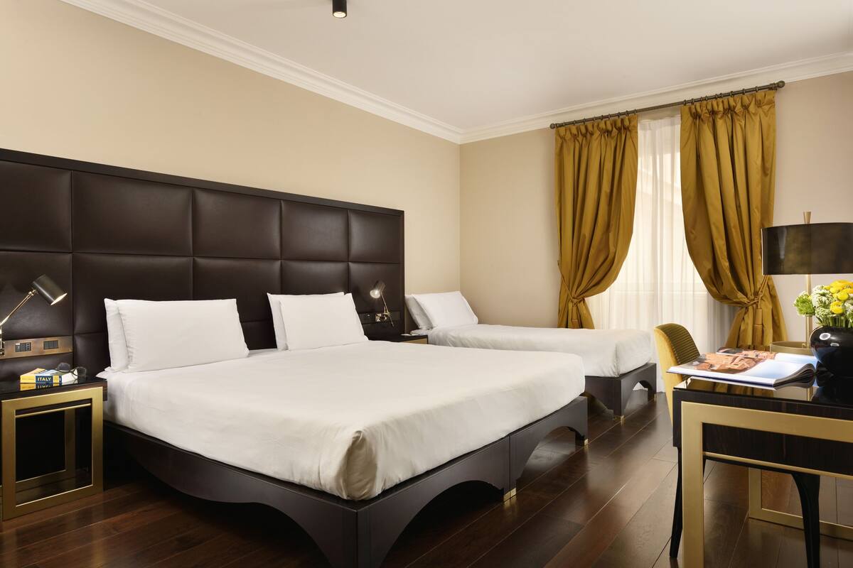 A hotel room with a large bed, a smaller bed, a desk with a lamp, and a view of the outdoors through curtains.