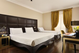 A hotel room with a large bed, a smaller bed, a desk with a lamp, and a view of the outdoors through curtains.