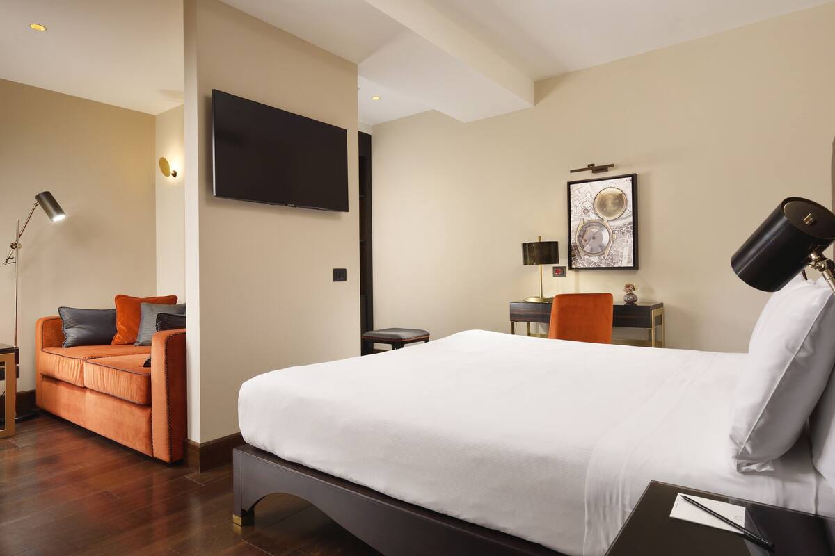 A hotel room with a bed, a sofa, a desk, a chair, a TV, and a lamp.