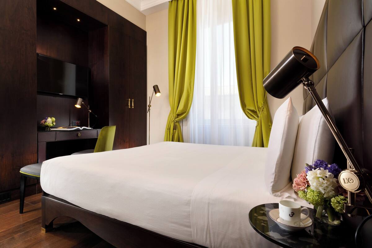 A hotel room with a large bed, a desk with a chair, a TV mounted on the wall, and a bedside table with a cup and flowers.