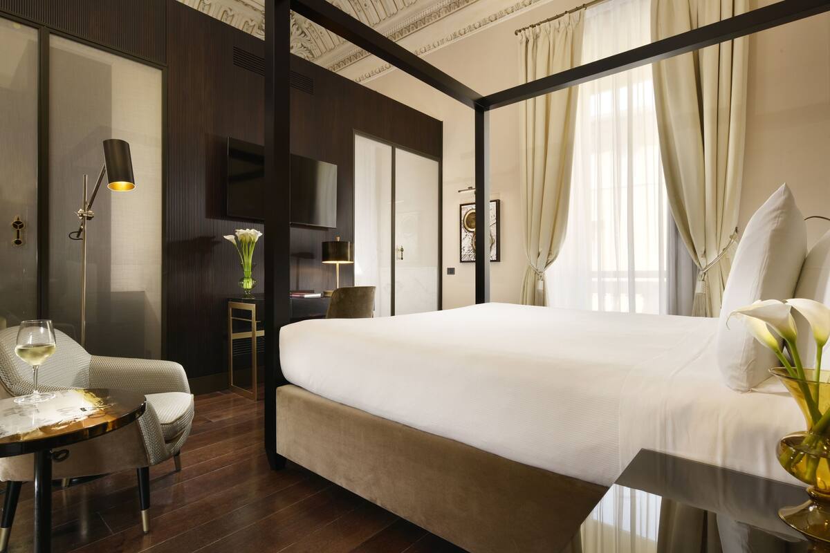 A modern hotel room with a large bed, a sitting area with a table and chair, a desk, and a large window with curtains.