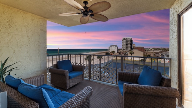 A balcony with wicker furniture and a ceiling fan, overlooking a beach and cityscape.