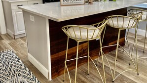 A kitchen with white cabinets, a stainless steel refrigerator, a built-in microwave, and a coffee maker. There are two gold bar stools and a sign that reads "Our Kitchen."