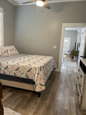 Un dormitorio con cama, suelo de madera, ventilador de techo y un pasillo que conduce a un baño.