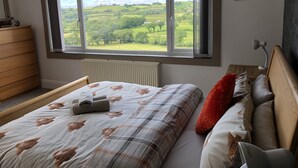 Une chambre avec une grande fenêtre, un lit recouvert d’une couette à motifs floraux, un radiateur, une commode en bois et un élément décoratif mural.