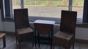 Une pièce avec deux fauteuils en osier et une petite table, de grandes fenêtres donnant sur un paysage de champs verdoyants et de collines.