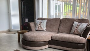 Un salon moderne avec un canapé d’angle, une grande fenêtre avec des stores, un tapis et un pouf en cuir.