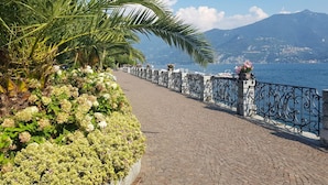 Ein gepflasterter Weg mit schmiedeeisernem Geländer, gesäumt von üppiger Vegetation und Blumen, führt zu einem Gewässer mit Bergen im Hintergrund.