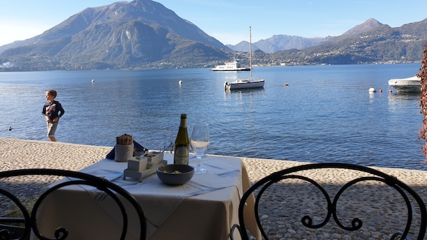 Ein Tisch am See mit einer Flasche und einem Glas, umgeben von Stühlen, mit Blick auf einen ruhigen See mit Booten und Bergen im Hintergrund.
