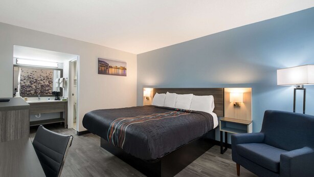 Quarto de hotel com cama, escrivaninha, cadeira, mesa de cabeceira e abajur.