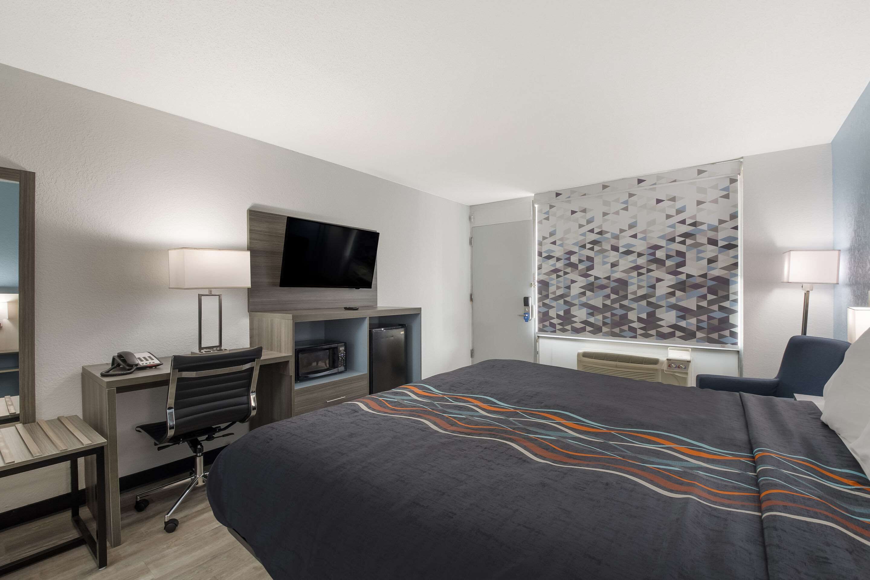 A hotel room with a bed, desk, chair, TV, and a patterned window treatment.