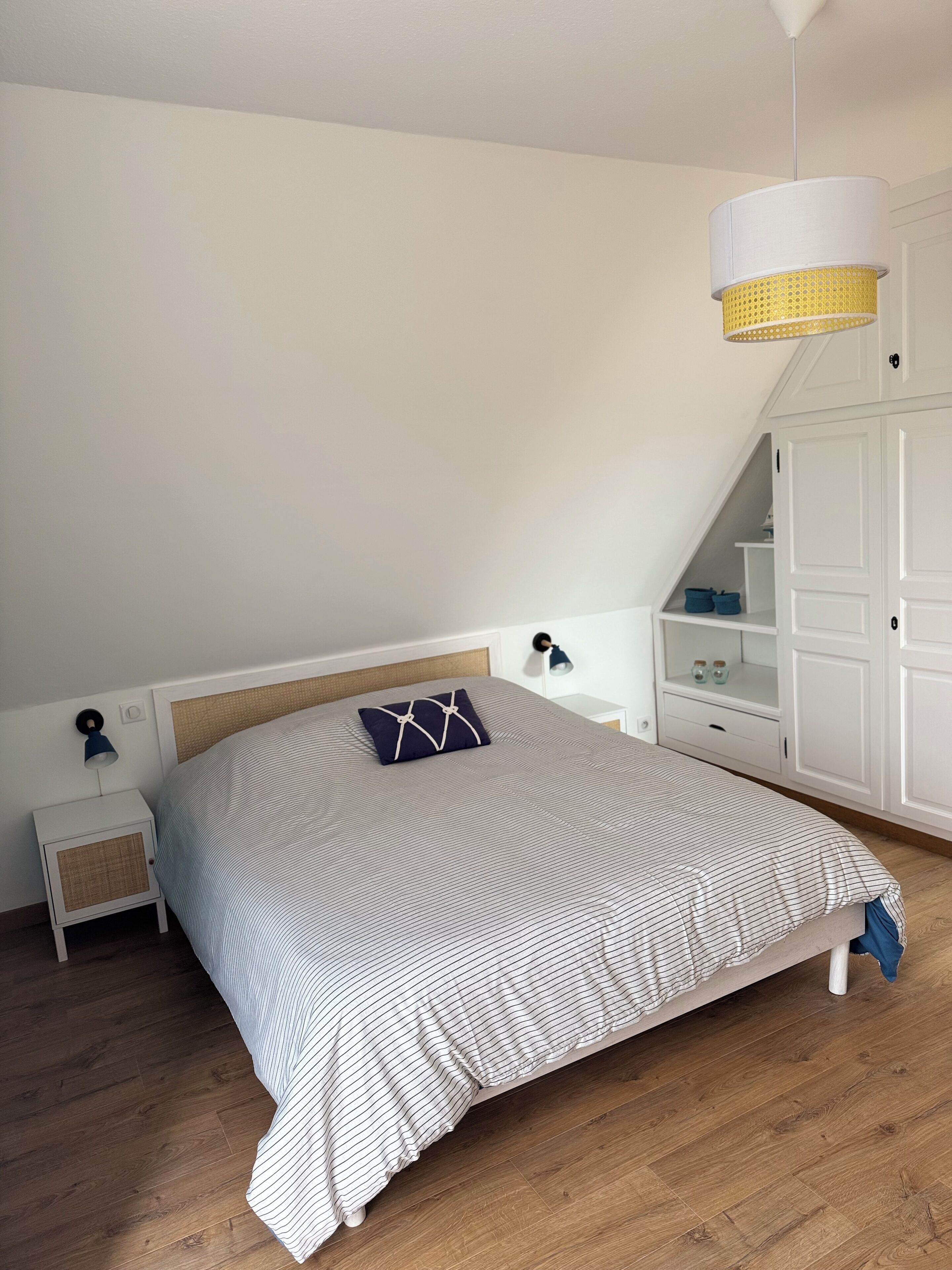 A bedroom with a bed, a nightstand, a lamp, a wardrobe, and a hanging light fixture.