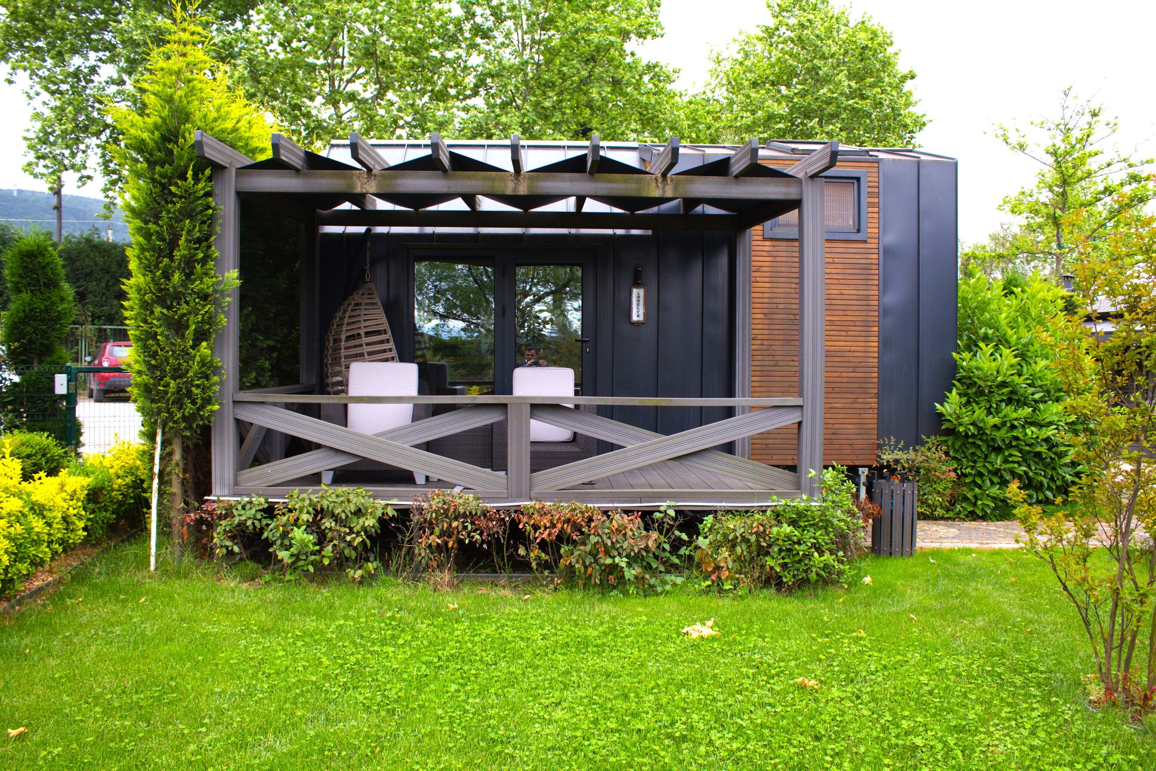 Un área de estar moderna al aire libre con terraza de madera y sillas, rodeada de abundante follaje y un edificio de fachada oscura.