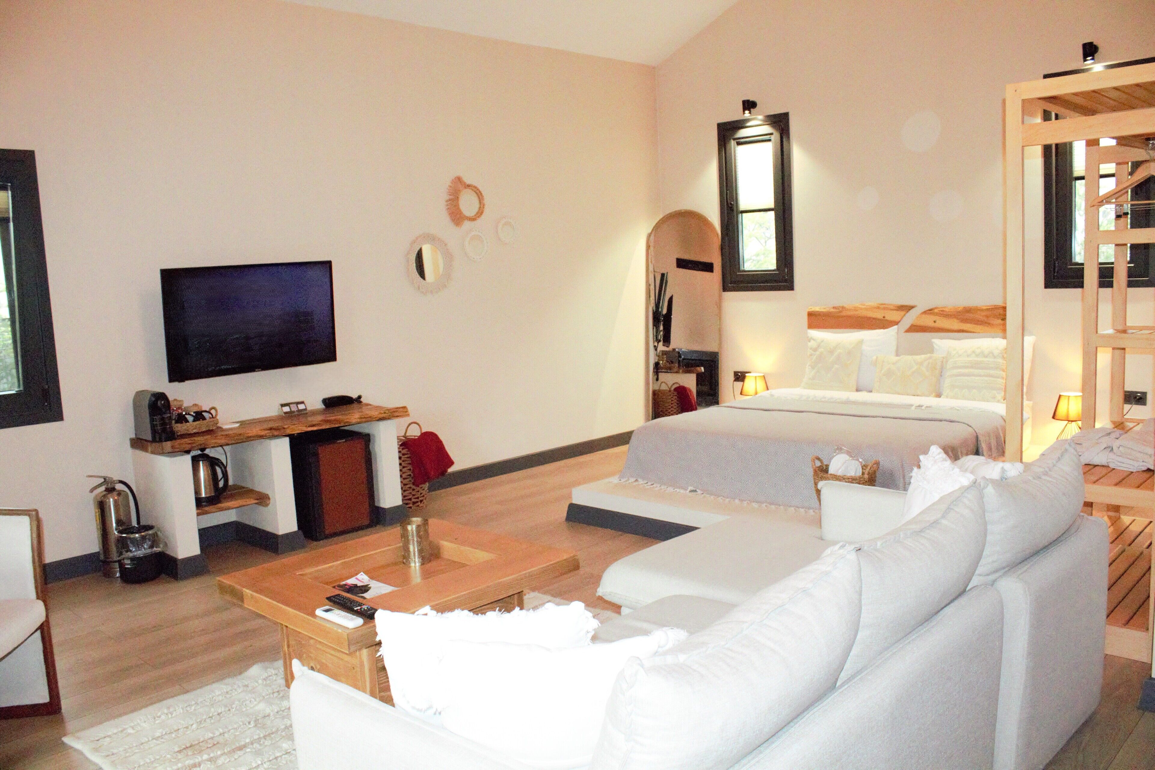 A modern hotel room with a sofa, a bed, a TV, a wooden coffee table, and a small kitchen area.