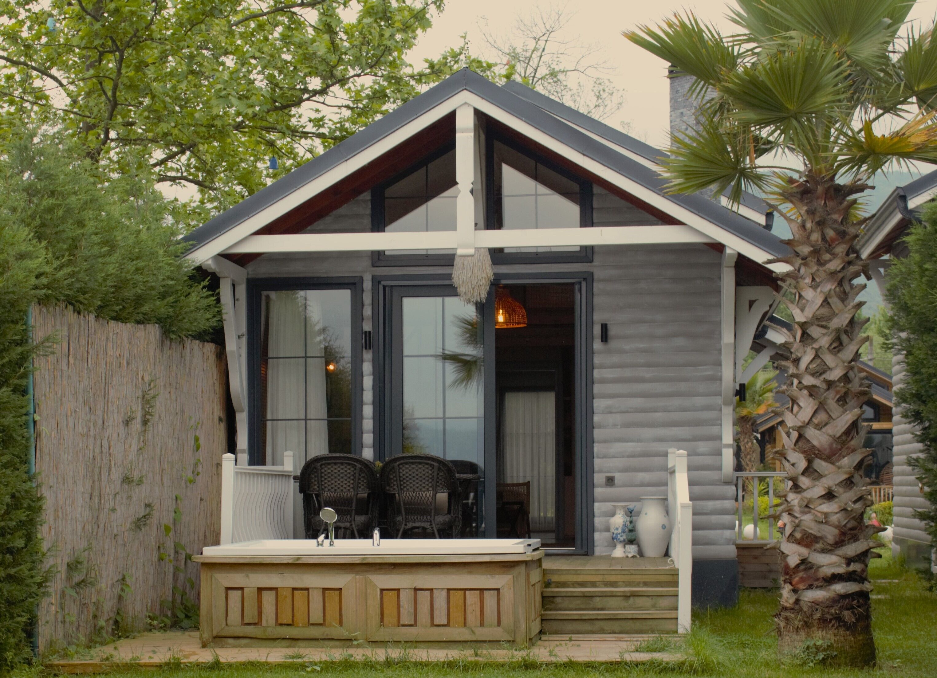Une maison moderne dotée d’une grande porte vitrée, d’une terrasse en bois avec deux chaises et d’un palmier.