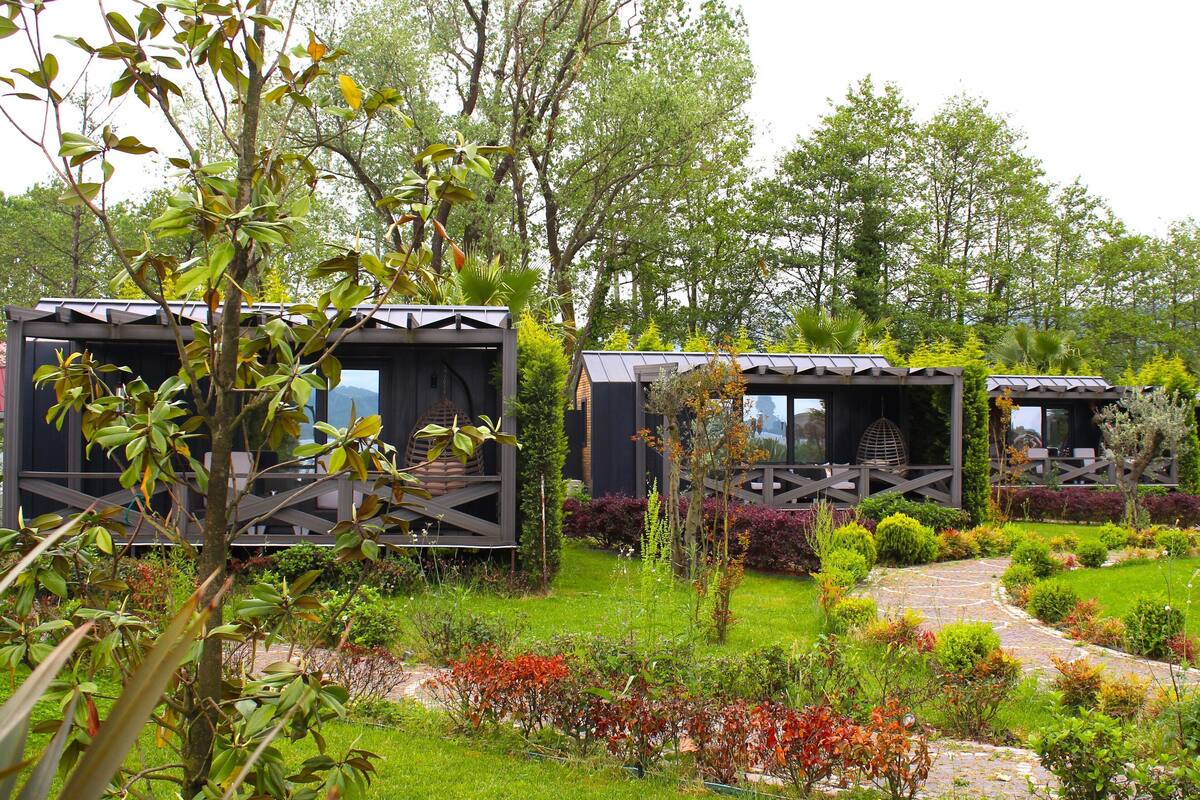 A garden with three wooden cabins, surrounded by lush greenery and a stone pathway.