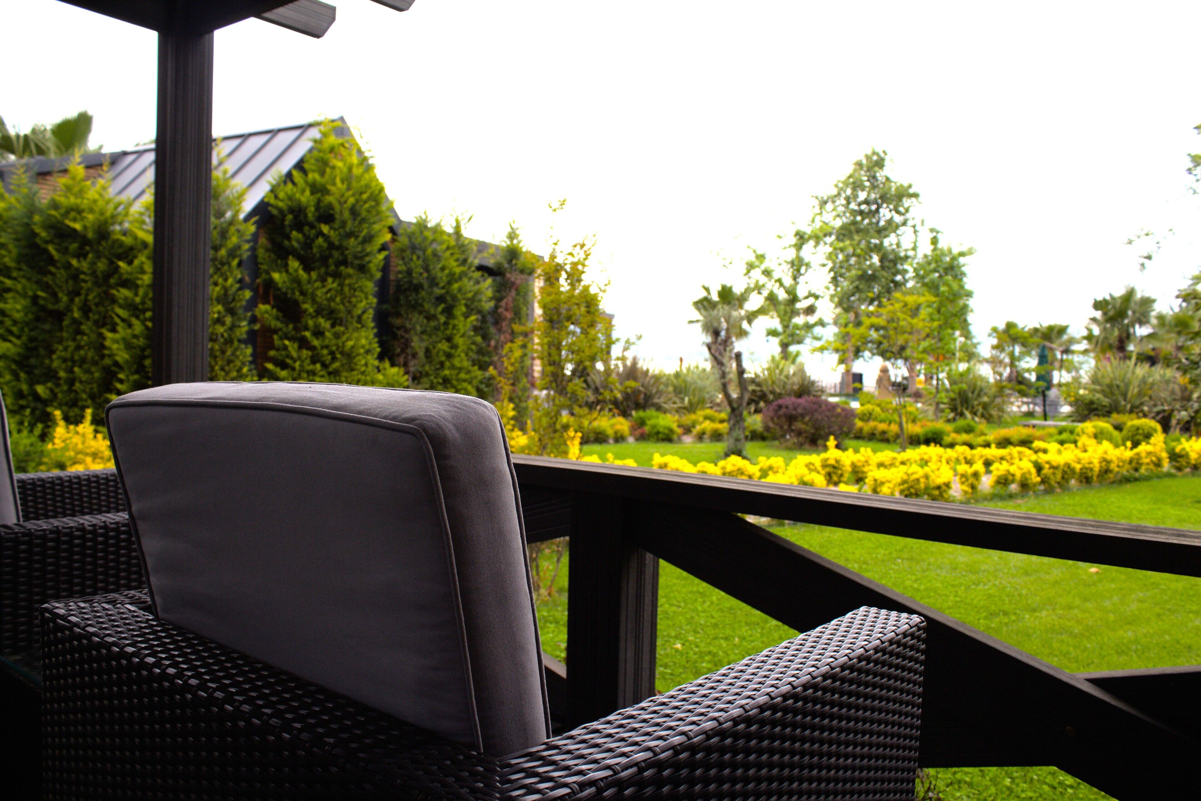 A wicker chair with a cushion, a wooden deck, and a view of a well-maintained garden with various plants and a lawn.