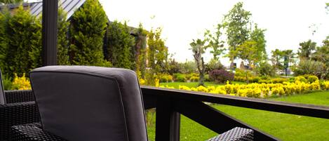 A wicker chair with a cushion, a wooden deck, and a view of a well-maintained garden with various plants and a lawn.