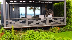 A wooden deck with two white cushioned chairs and a wicker chair, overlooking a lush garden and a view of the ocean. - Gölevi Resort Sapanca (Sapanca)