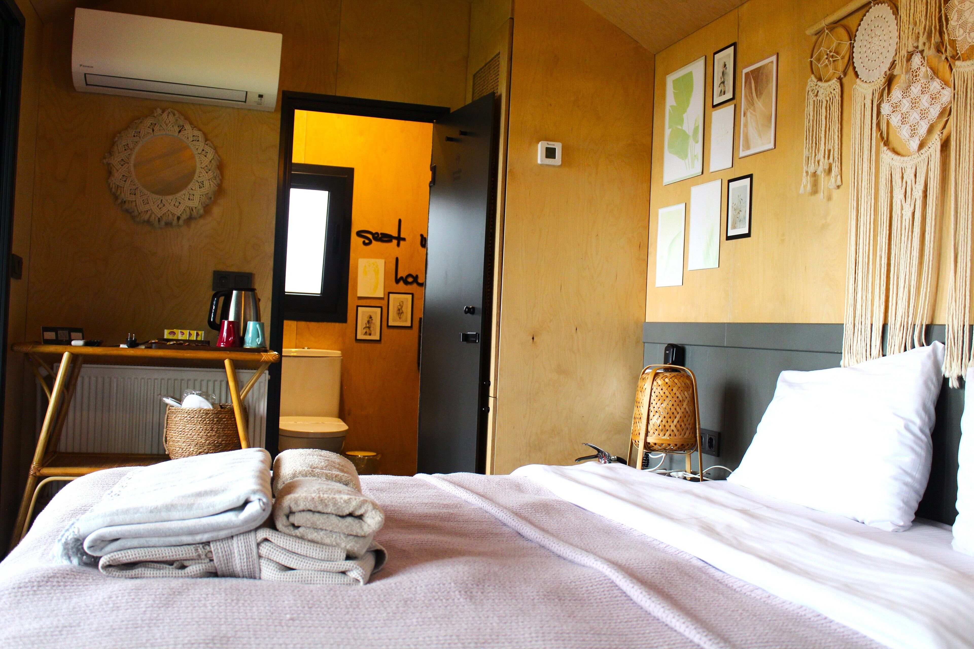 A hotel room with a bed, a bedside table, a wall-mounted air conditioner, a mirror, and a door leading to another room.