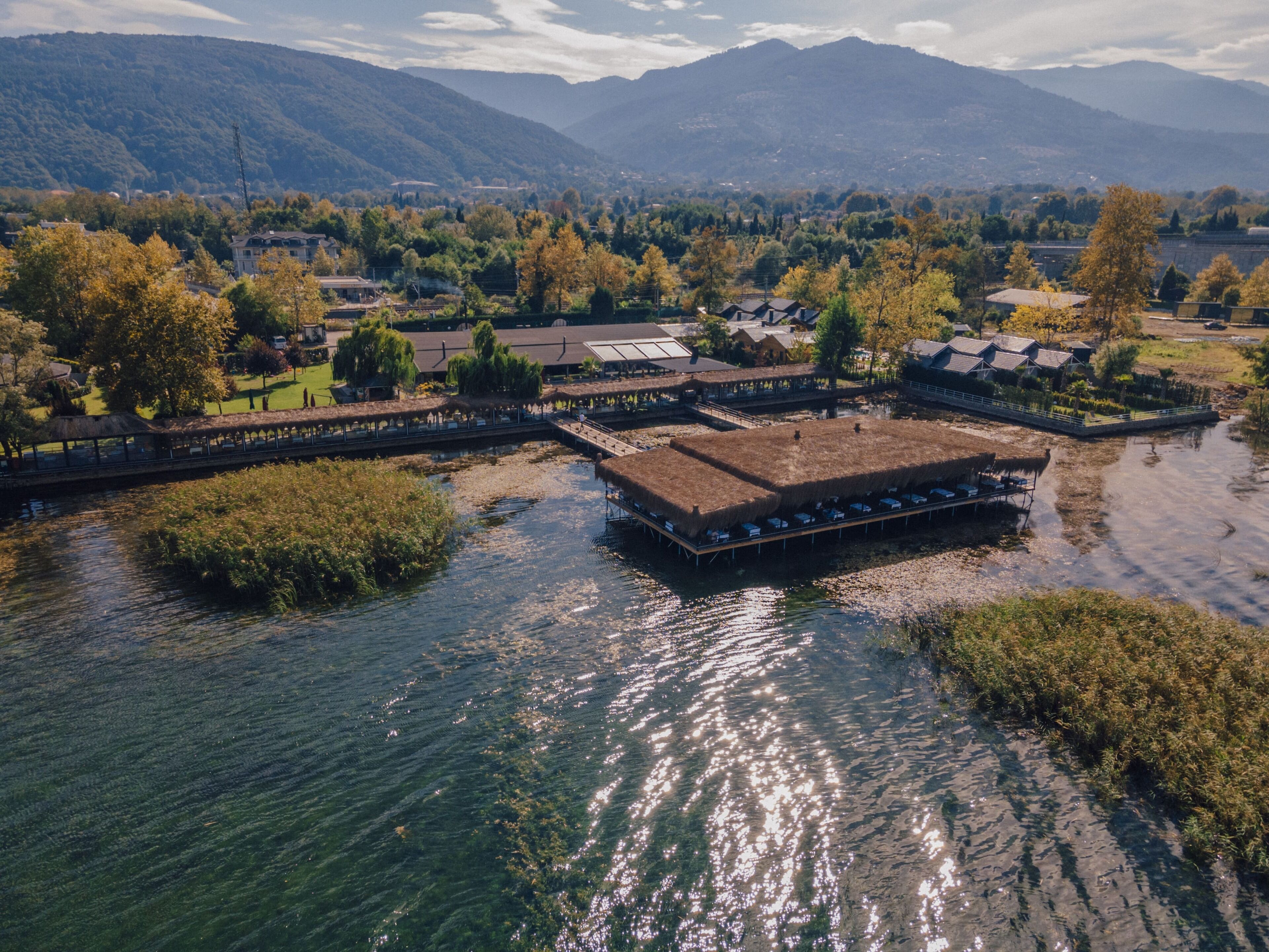 An aerial view of a riverside resort with thatched-roof buildings, a wooden dock, and a backdrop of mountains.