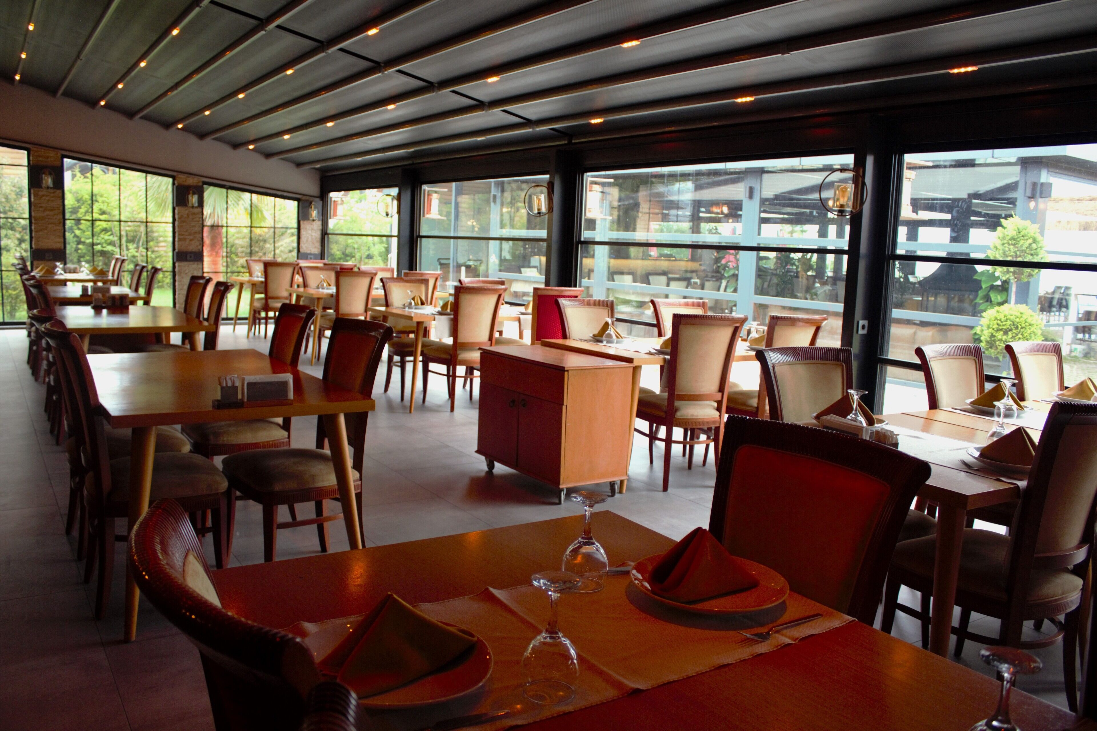 A spacious restaurant with multiple tables and chairs, large windows offering a view of the outdoors, and a modern ceiling design.